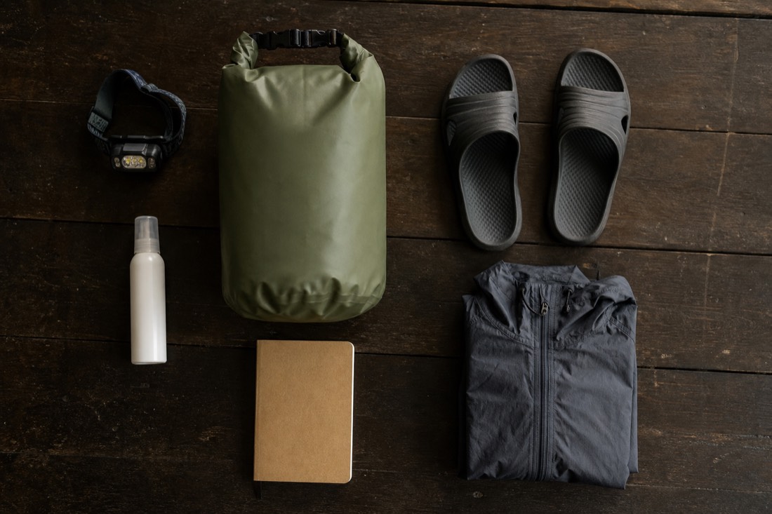 Flat lay of jungle retreat gear: headlamp, dry bag, insect repellent, sandals, journal and rain jacket