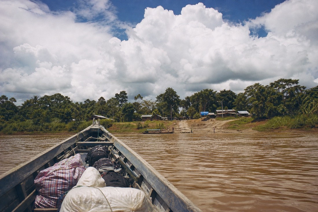 Long wooden riverboat loaded with bags approaching a muddy Amazon landing under big clouds