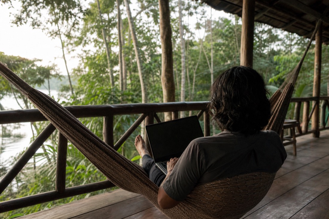Traveler seen from behind reading a laptop in a hammock on a jungle lodge porch