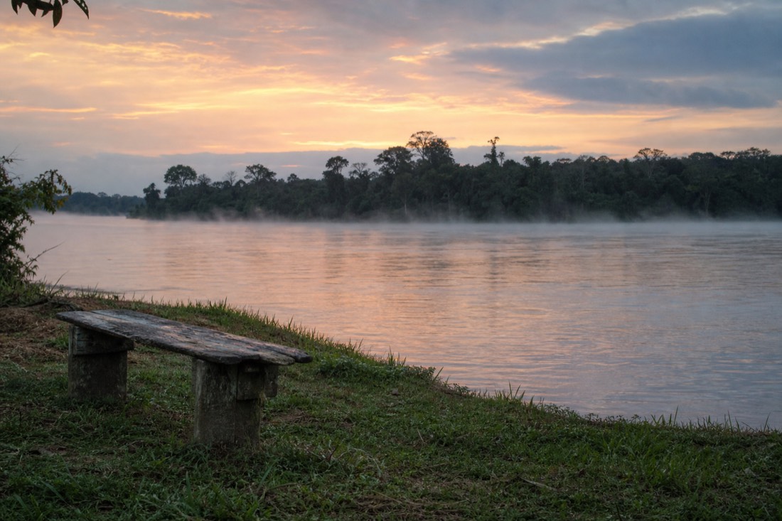 Calm wide river at first light with low mist and a single empty wooden bench on the bank, Amazon river near Tamshiyacu