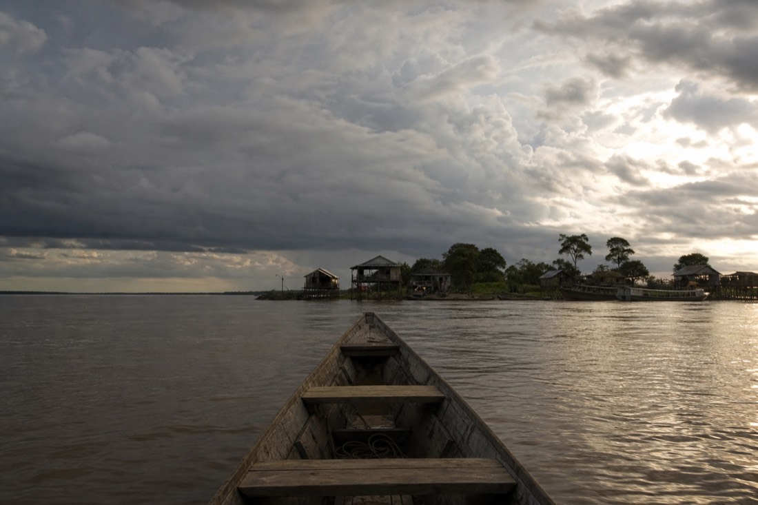 Long wooden boat on the Amazon river approaching a small jungle village dock, dramatic clouds, Loreto region