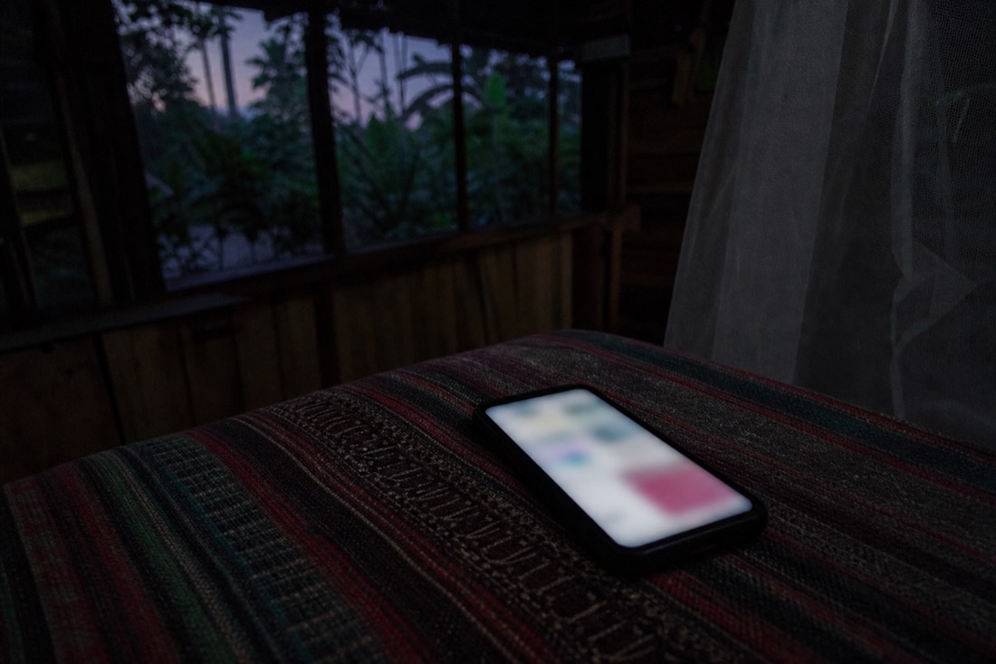Glowing phone screen lying on a woven blanket in low evening light inside a wooden jungle room, screen content blurred, Loreto region