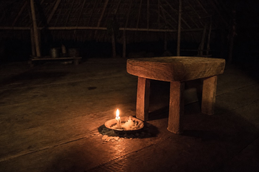 Small candle burning low beside a wooden stool inside a dark maloca, wax pooled on the boards