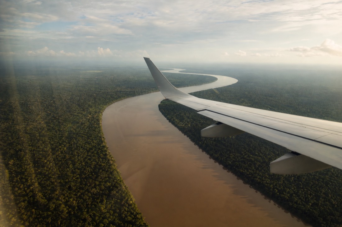 Airplane wing over unbroken green Amazon rainforest with a winding brown river below