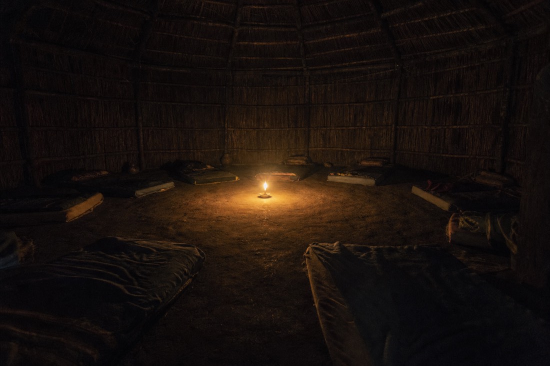 Dim interior of a thatched maloca at night lit by a single candle, empty mattresses along the walls