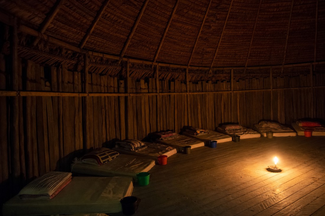 Row of thin mattresses with folded blankets and small buckets along the curved wall of a candlelit maloca