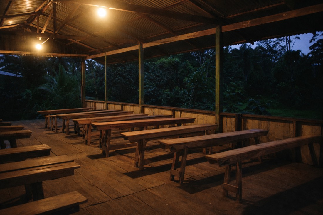 Rows of simple wooden benches in an open-sided community hall at dusk, warm lamplight, Iquitos region