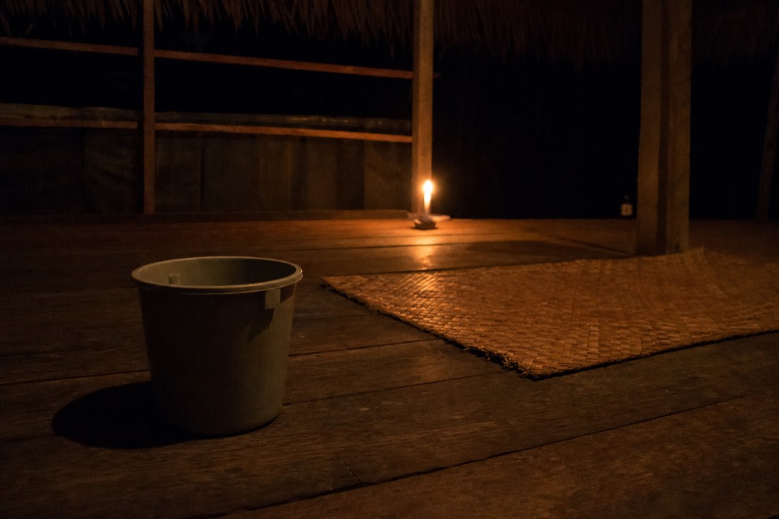 Plastic bucket beside a woven mat on the wooden floor of a candlelit maloca at night
