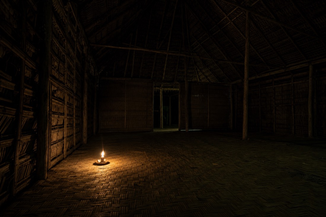 Single low candle burning inside a dark empty maloca with woven mats in shadow